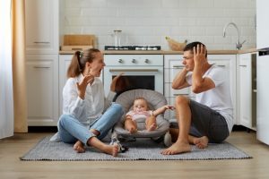 Indoor Short Of Arguing Couple Sitting On The Floor In Kitchen Wife Screaming Loudly Husband Covering Ears With Palms Family Posing With Infant Baby In Rocking Chair 1